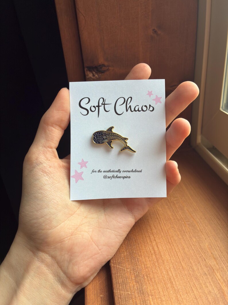 Pu&ograve; includere: Una spilla in smalto a forma di squalo balena dorato con dettagli neri &egrave; esposta su una carta bianca. La carta recita "Soft Chaos" in corsivo nero, con accenti di stelle rosa. &Egrave; presente anche il testo "for the aesthetically overwhelmed @softchaospins".