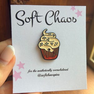 May include: An enamel pin featuring a cupcake with a snake-shaped cream swirl, decorated with colorful sprinkles, attached to a white display card with the text Soft Chaos, for the aesthetically overwhelmed, and @softchaospins.
