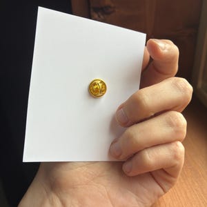 Pu&ograve; includere: Una piccola spilla rotonda color oro &egrave; centrata su una carta bianca. La spilla ha una superficie strutturata. La carta &egrave; tenuta in mano, con una superficie di legno sullo sfondo.