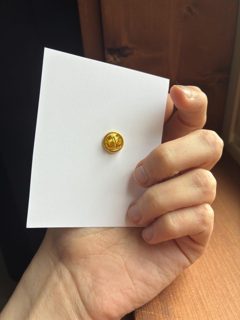 Pu&ograve; includere: Una piccola spilla rotonda color oro &egrave; centrata su una carta bianca. La spilla ha una superficie strutturata. La carta &egrave; tenuta in mano, con una superficie di legno sullo sfondo.