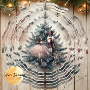 May include: A decorative, layered metal ornament featuring a Christmas scene with a ballerina, nutcracker, and Christmas tree. The ornament has a circular shape with a wavy, layered design and the text "Lunari Designs". The color palette includes silver, pink, and green.