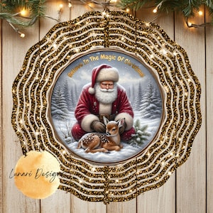 Peut inclure: Un carillon à vent circulaire en métal avec une finition à paillettes noires et dorées, illustrant le Père Noël agenouillé dans une forêt enneigée avec un faon. Le texte Believe In The Magic Of Christmas est imprimé en doré au-dessus.