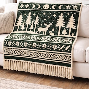 Peut inclure: Une couverture tricotée vert foncé et crème avec un motif de forêt et de lune. La couverture présente un cerf, des arbres et des motifs géométriques, avec des franges crème le long du bord inférieur. Elle est drapée sur un canapé blanc.