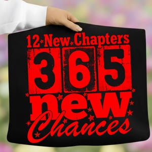 May include: Black pillowcase with the text "12 New Chapters 365 New Chances" in red. The numbers and words are in a distressed font, with star accents. The pillowcase is held by a person.