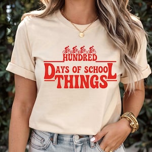 May include: A beige t-shirt with rolled sleeves, displaying red text and graphics. The text reads "Hundred Days of School Things" with a bicycle graphic above. The shirt is made of a soft material.