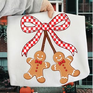 Puede incluir: Un artículo de tela blanca con un diseño festivo. El diseño incluye dos hombres de jengibre con pajaritas rojas, suspendidos de un lazo a cuadros rojo y blanco. Los hombres de jengibre tienen caras sonrientes y son de color marrón.