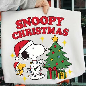 Op de afbeelding: Witte draagtas met Snoopy in een kerstmuts die een kerstboom versiert. De tekst "SNOOPY CHRISTMAS" is rood. Er is ook een kleine gele vogel aanwezig. Cadeaus liggen onder de boom.