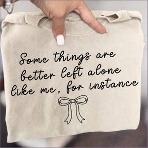 May include: A beige sweatshirt with the text "Some things are better left alone like me, for instance" in black script. A bow graphic is below the text. The sweatshirt is folded.