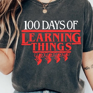 May include: Dark grey t-shirt featuring the text "100 DAYS OF LEARNING THINGS" in white and red. The design includes red upside-down bicycles. The shirt has a vintage, worn appearance.