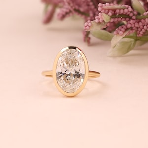 Puede incluir: Un anillo de oro con un diamante de talla ovalada. El diamante está engastado en un bisel, y la banda es de oro liso. El anillo está sobre una superficie rosa claro, con flores rosas en el fondo. Dimensiones del diamante: 1,5 cm x 1 cm.