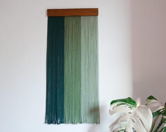 May include: A wall hanging with a wooden top and vertical strands of yarn in shades of green. The yarn is arranged in three vertical sections, transitioning from dark teal to light green. The piece is mounted on a white wall.