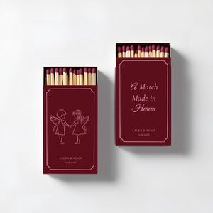 May include: Two burgundy matchboxes with white text and illustrations. One box reads "A Match Made in Heaven" and the other features two angel figures. Both boxes have the names "LAURA & ADAM" and a date.