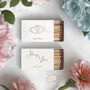 Può includere: Due scatole di fiammiferi bianche con cassetti aperti che rivelano fiammiferi di legno. Una scatola presenta un disegno di due cuori intrecciati e i nomi "Emily & Michael". L'altra scatola recita "Always Yours" e la data "16.05.2027."