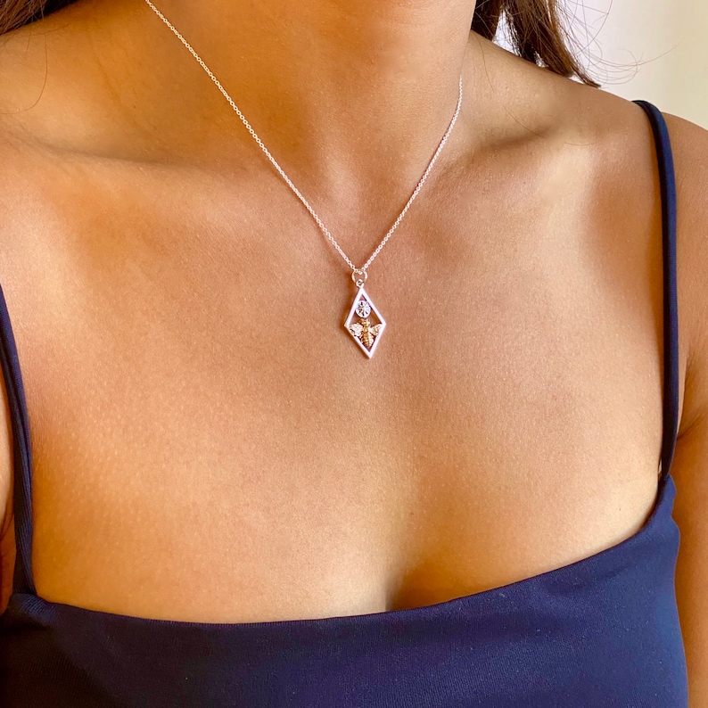May include: A silver necklace with a diamond-shaped pendant. The pendant features a geometric design with a bee and flower motif. The necklace is worn on a person wearing a navy blue top.