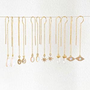 Può includere: Una collezione di orecchini pendenti dorati con vari ciondoli. I ciondoli includono opali, cristalli trasparenti e disegni a stella. Gli orecchini pendono da delicate catene, creando un effetto pendente. Sono esposti su sfondo bianco.
