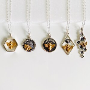May include: Five silver necklaces with different pendant designs. The pendants include a honeycomb with a bee, a sunflower with a bee, a circular pendant with a bee and moon phases, a diamond-shaped pendant with a bee and a star, and a floral pendant with a bee.