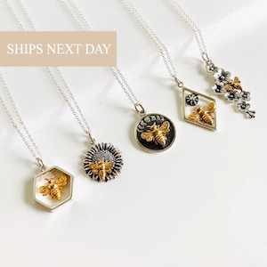 May include: A collection of silver necklaces with various pendant designs. The pendants include a gold-colored bee inside a hexagon, a sunflower with a bee, a circular pendant with a bee and moon phases, a diamond-shaped pendant with a bee, and a floral design.