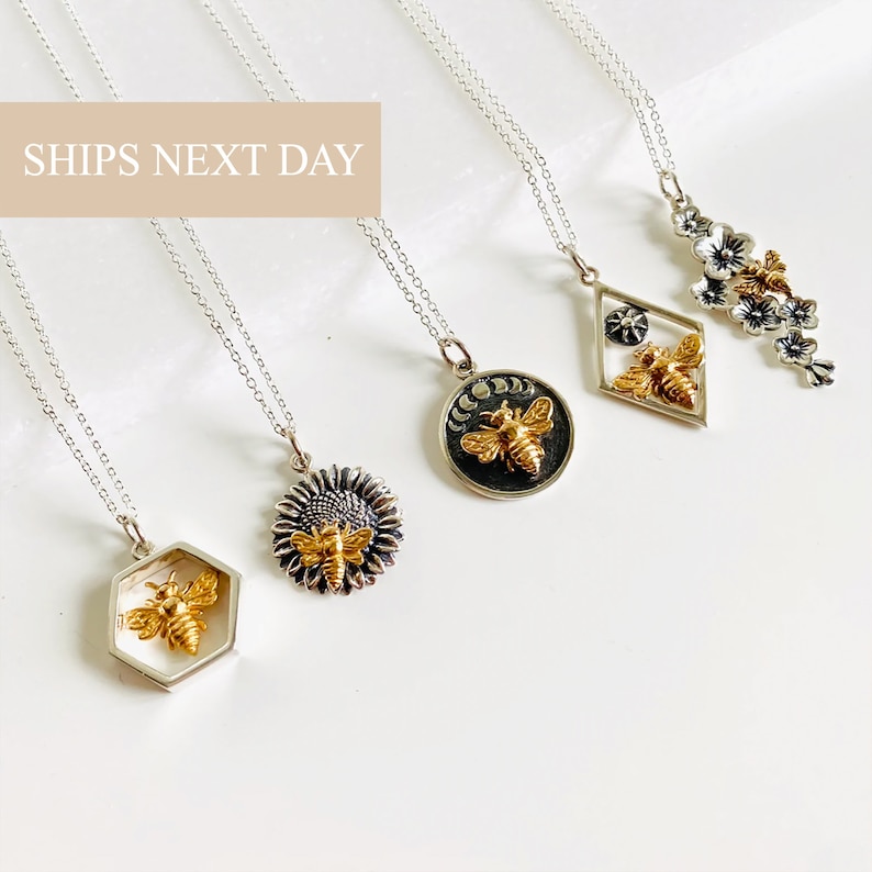 May include: A collection of silver necklaces with various pendant designs. The pendants include a gold-colored bee inside a hexagon, a sunflower with a bee, a circular pendant with a bee and moon phases, a diamond-shaped pendant with a bee, and a floral design.