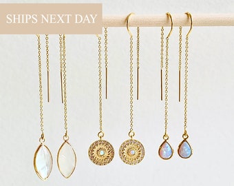 May include: A collection of gold-colored threader earrings with various pendant designs. The earrings are displayed on a wooden dowel against a white background. The pendants include teardrop-shaped opals, circular designs with small stones, and elongated, oval-shaped pendants.