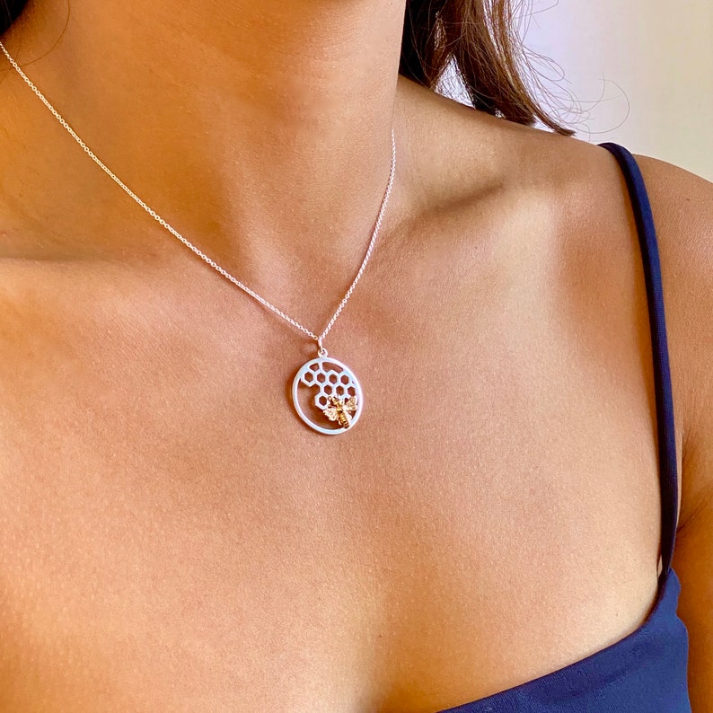May include: Silver necklace with a circular pendant featuring a honeycomb design and a gold-colored bee. The pendant hangs from a delicate silver chain. The necklace is worn against a person's skin.
