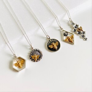 May include: A collection of silver necklaces with various pendant designs. Each pendant features a gold-colored bee. Designs include a hexagon, sunflower, moon phases, a diamond shape with a star, and a floral branch.