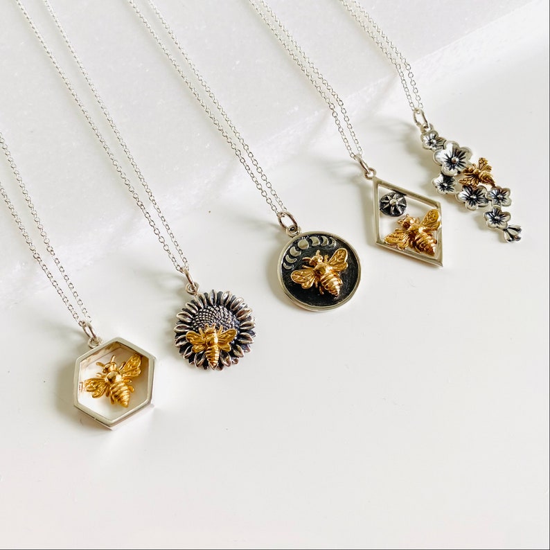May include: A collection of silver necklaces with various pendant designs. Each pendant features a gold-colored bee. Designs include a hexagon, sunflower, moon phases, a diamond shape with a star, and a floral branch.