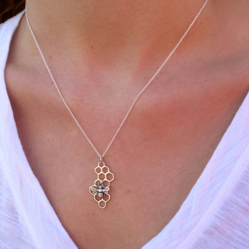 May include: A silver necklace with a bee and honeycomb pendant. The necklace is on a model wearing a white shirt.