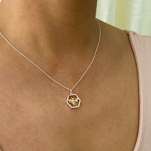 May include: A silver necklace with a hexagonal pendant featuring a gold-colored bee design. The bee is centered within the pendant, which is suspended from a delicate silver chain. The necklace is worn against a light pink top.