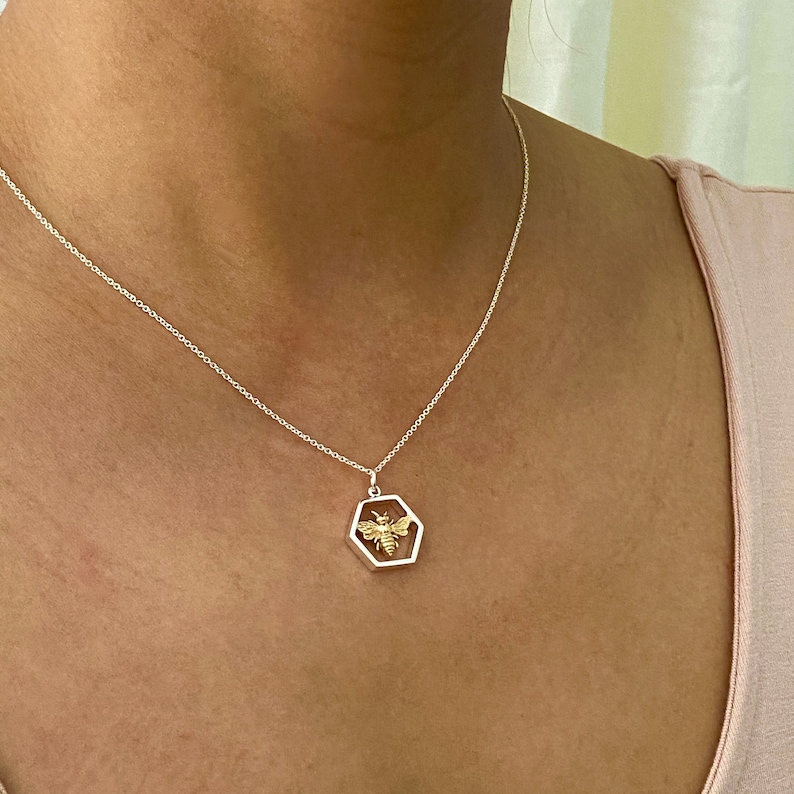 May include: A silver necklace with a hexagonal pendant featuring a gold-colored bee design. The bee is centered within the pendant, which is suspended from a delicate silver chain. The necklace is worn against a light pink top.