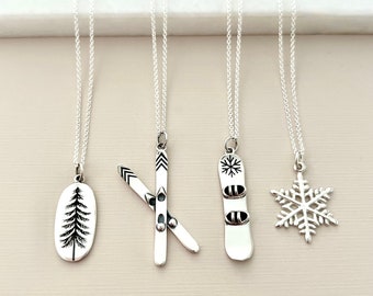 Puede incluir: Cuatro collares de plata con diferentes colgantes de temática invernal: un pino, un par de esquís, una tabla de snowboard y un copo de nieve.