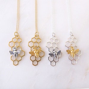 May include: Three gold and silver necklaces with a honeycomb pendant and a small bee charm. The necklaces are on a white background.