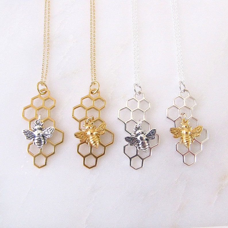 May include: Three gold and silver necklaces with a honeycomb pendant and a small bee charm. The necklaces are on a white background.