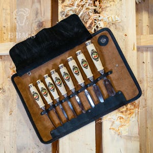 May include: A set of wood carving chisels in a brown leather roll. The chisels have light-colored handles with a cherry logo and silver metal accents. The roll has a black suede flap.