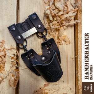 May include: Black leather hammer holder with a metal accent and rivets. The holder has a loop for attaching to a belt or other item. The text "HAMMERHALTER KARABINER" is visible on the side. The holder is designed to carry a hammer.