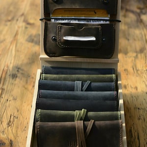 May include: A wooden display case holding several leather pouches and wallets. The top section features a dark brown leather pouch with a brass emblem. The lower section displays various rolled-up leather items in shades of green, blue, and brown.
