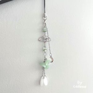 Puede incluir: Un charm de teléfono plateado con un cordón negro, con un charm en forma de nube, cuentas verdes, una luna creciente y dijes blancos en forma de lágrima. El charm está sujeto a un teléfono.