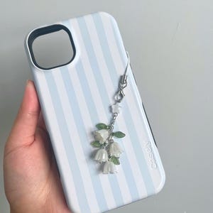 May include: A light blue and white striped phone case with a silver charm featuring white flower and green leaf accents. The case has a black camera cutout and the brand name "Casely" is printed on the side.
