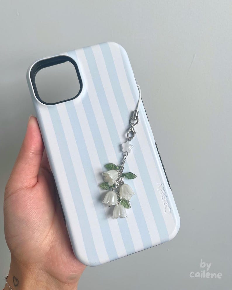 May include: A light blue and white striped phone case with a silver charm featuring white flower and green leaf accents. The case has a black camera cutout and the brand name "Casely" is printed on the side.