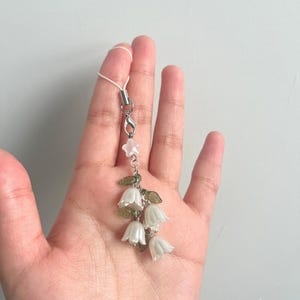 May include: A delicate floral charm featuring white bell-shaped flowers, green leaves, and a star-shaped accent. The charm is attached to a silver-colored clasp and a white cord. The design is reminiscent of lily of the valley.