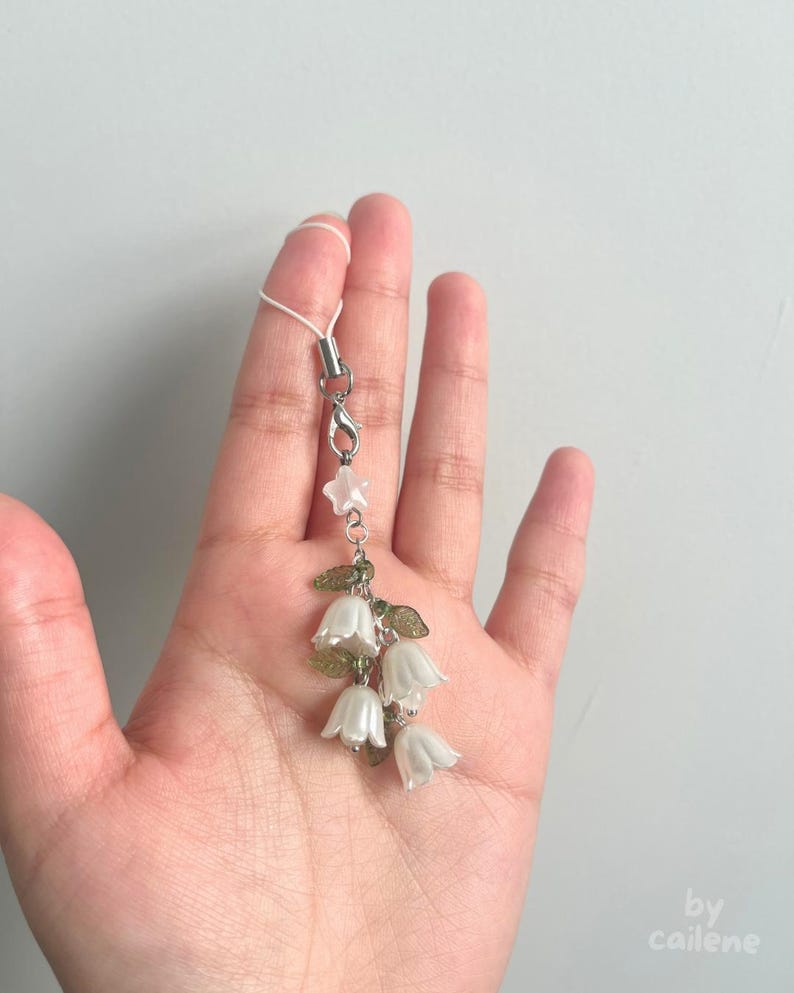 May include: A delicate floral charm featuring white bell-shaped flowers, green leaves, and a star-shaped accent. The charm is attached to a silver-colored clasp and a white cord. The design is reminiscent of lily of the valley.