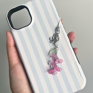 May include: A white and light blue striped phone case with a floral charm. The charm features pink flower beads, a silver flower, a green leaf, and a white bead. The phone case has the word "Casely" printed on it.