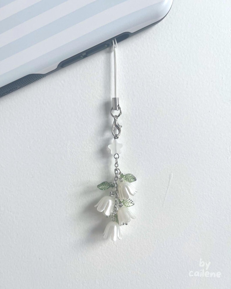 May include: A white phone charm featuring a cluster of white flower blossoms with green leaves and a star-shaped bead. The charm is attached to a silver-colored clasp and a white cord, hanging from a phone.