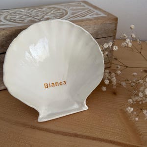 Puede incluir: Un plato de cerámica blanco brillante con forma de concha de vieira. La superficie presenta un acabado liso y reflectante con el nombre Bianca impreso en letras doradas cerca del centro de la concha, resaltando su diseño curvo y estriado.