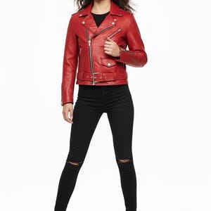 May include: A vibrant red leather jacket featuring a belted waist and silver zips. The jacket is worn with black skinny jeans with knee rips and black ankle boots. The model is standing against a white backdrop.
