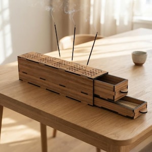 Puede incluir: Un quemador de incienso rectangular de madera con acabado marrón oscuro, que presenta una parte superior perforada para tres varitas de incienso y dos cajones extraíbles en el lateral para almacenamiento. La caja tiene detalles de unión visibles.