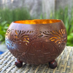 Puede incluir: Un cuenco artesanal de cáscara de coco marrón con intrincadas tallas de una tortuga y motivos florales. El cuenco tiene un interior dorado y descansa sobre tres pequeños pies de color marrón oscuro. El diseño incluye detalles recortados.