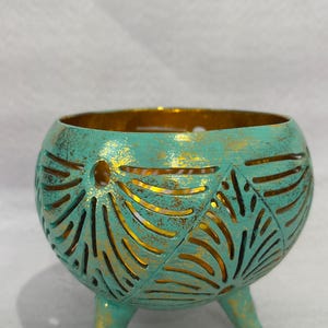 Könnte beinhalten: Ein teal-farbener, runder Kerzenhalter mit einem Used-Look-Finish und goldenen Akzenten. Der Metallhalter hat blattförmige Aussparungen und drei kleine Beine. Der Innenraum hat eine goldene Oberfläche und erzeugt ein warmes Leuchten, wenn er beleuchtet wird.