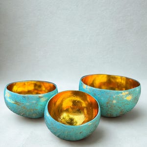 May include: Three decorative bowls with a turquoise exterior and a gold interior. The bowls have a distressed finish, giving them a rustic appearance. The bowls are arranged in a group, showcasing their varying sizes.