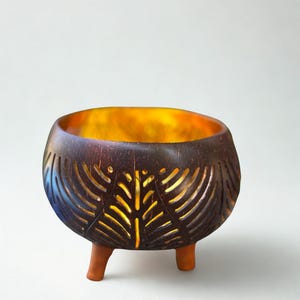 May include: A handcrafted, dark brown candle holder with a leaf-like pattern carved into the sides. The interior glows with a warm, golden light. The holder is supported by three small, light brown legs, creating a unique decorative piece.