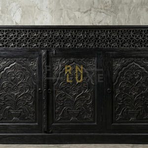May include: Black wooden cabinet with detailed floral carvings on the doors and an openwork design at the top. The cabinet has three doors and is flanked by large terracotta pots with green plants.